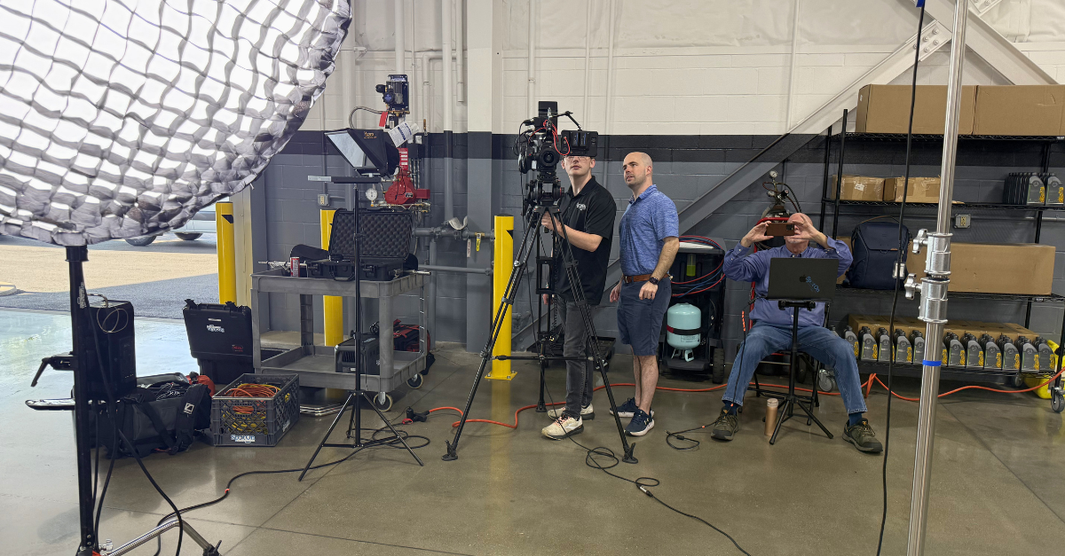 Film crew setting up video production shoot in warehouse studio with camera, tripod, softbox light, and monitor equipment.